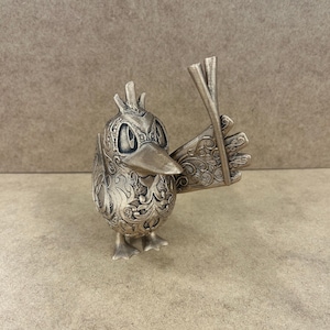 May include: A silver bird figurine with detailed floral patterns. The bird holds chopsticks in its wing, and has a detailed face with a beak and eyes. The figurine is standing on two feet.