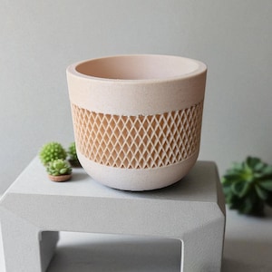 Może przedstawiać: Jasnoróżowa ceramiczna doniczka z geometrycznym wzorem kratki pośrodku. Doniczka stoi na małym, szarym betonowym stojaku, z małymi sukulentami w pobliżu. Do użytku wewnątrz i na zewnątrz.