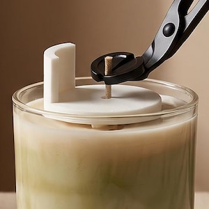 May include: A clear glass candle container filled with marbled green and white wax. A white circular wick centring tool sits on top of the wax, surrounding a single wooden wick. A pair of black metal wick trimmers is positioned above the wick.