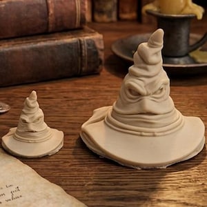 May include: Two cream-colored, pointed, wide-brimmed hat figurines with facial features resembling eyes and a mouth, placed on a wooden surface alongside a piece of parchment with printed text, metal coins, a lit candle, and an hourglass.