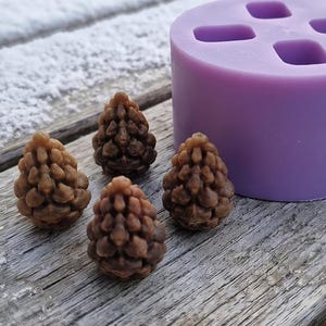 Puede incluir: Un molde de silicona morado de forma cilíndrica con cuatro cavidades en forma de piñas, acompañado de cuatro pequeños objetos marrones texturizados con forma de piña moldeados, que muestran detalles intrincados en sus escamas.