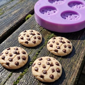 Könnte beinhalten: Eine runde, violette Silikonform mit vier kreisförmigen Vertiefungen und drei kleinen, runden, beigefarbenen Keksen mit dunkelbraunen Schokoladenstückchen. Die Kekse haben eine strukturierte Oberfläche und sind in einer Gruppe nahe der Form angeordnet.