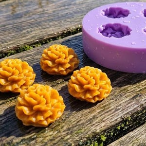 May include: A round, light purple silicone mold with four circular cavities, accompanied by four small, golden-yellow, flower-shaped wax or resin castings that feature intricate, layered petal details and a textured, ruffled appearance.