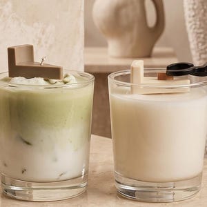 May include: Two glass candles in clear tumblers. The left candle features a layered green and white wax design topped with a brown L-shaped wax piece. The right candle contains solid white wax with a cream L-shaped wax piece and a black wick trimmer.