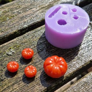 Peut inclure: Un moule en silicone violet avec diverses formes et tailles, à côté de quatre petits objets rouges en forme de tomate. Les objets sont disposés sur une surface en bois vieilli, suggérant un thème artisanal ou culinaire.