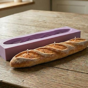 Pode incluir: Uma vela em forma de baguete com uma crosta dourada e três pavios brancos visíveis, posicionada em frente a um molde retangular de silicone roxo. A vela apresenta uma superfície texturizada que imita a aparência de um pão assado.