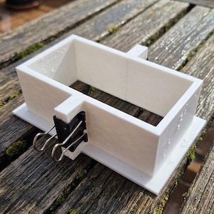 Può includere: Un telaio per stampo rettangolare bianco costruito con quattro pareti in plastica a incastro fissate su una piastra di base piatta. Una clip in metallo nero fissa uno dei giunti laterali del telaio, mantenendo la struttura salda.