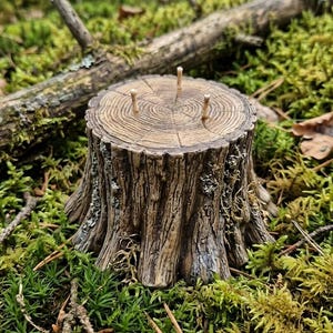 May include: A miniature tree stump with three small wooden pegs, set amidst green moss and forest debris. The stump is detailed with realistic bark texture and growth rings, creating a naturalistic aesthetic. The scene evokes a woodland theme.