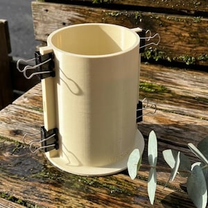 Op de afbeelding: Een crèmekleurige, cilindrische mal met zwarte bindclips. De mal staat op een houten oppervlak met groen. Waarschijnlijk voor knutsel- of doe-het-zelfprojecten. De clips houden de onderdelen van de mal vast.