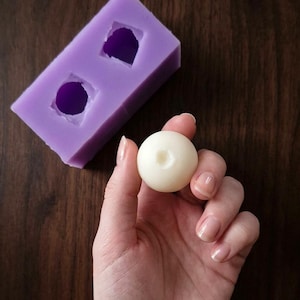 Peut inclure: Une sphère faite à la main, blanc cassé, tenue dans une main, à côté d'un moule en silicone violet avec deux cavités. Le moule a une forme rectangulaire avec une cavité arrondie et une en forme de losange. Fond en bois brun foncé.