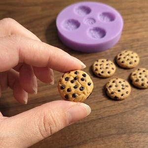 Op de afbeelding: Een miniatuur beige koekje met donkerbruine chocoladestukjes tussen de vingers, vergezeld door vier identieke miniatuurkoekjes en een ronde paarse siliconen mal met vijf cirkelvormige uitsparingen voor het knutselen.