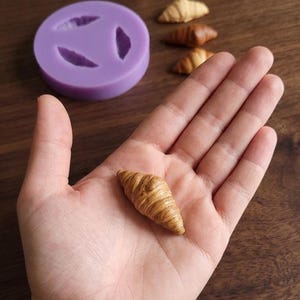 Op de afbeelding: Een gedetailleerde miniatuur croissant in een hand, met realistische kleuring en textuur. Een paarse siliconen mal met croissantvormige holtes staat op de achtergrond, samen met andere miniatuur croissants.