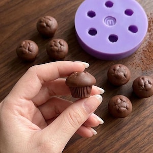 Op de afbeelding: Een miniatuur chocolademuffin wordt tussen vingers gehouden, met meerdere muffins en een paarse siliconen mal op de achtergrond. De muffins hebben een gedetailleerd, getextureerd uiterlijk, wat wijst op een bak- of knutselthema.