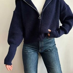 May include: A navy blue zip-up sweater with a hood and long sleeves. The sweater is paired with blue jeans. The sweater has a ribbed texture and a silver zipper. The model is wearing a white t-shirt underneath the sweater.