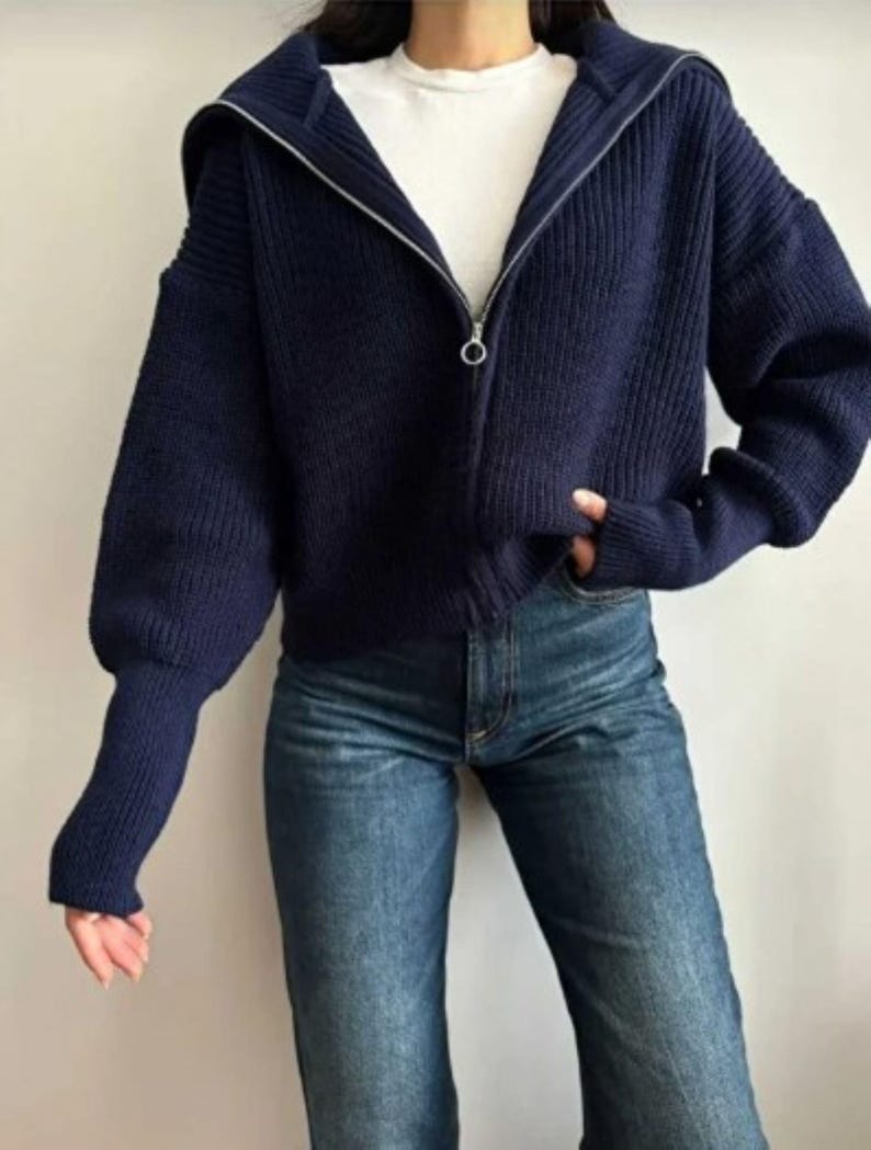 May include: A navy blue zip-up sweater with a hood and long sleeves. The sweater is paired with blue jeans. The sweater has a ribbed texture and a silver zipper. The model is wearing a white t-shirt underneath the sweater.