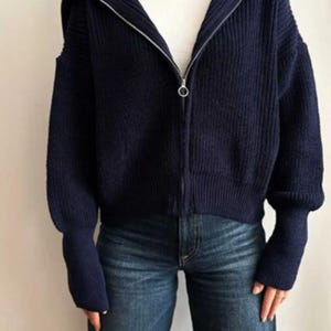 May include: A navy blue zip-up hooded sweater with a silver zipper. The sweater has a ribbed knit texture and long sleeves. The person is wearing blue jeans and a white t-shirt underneath the sweater.