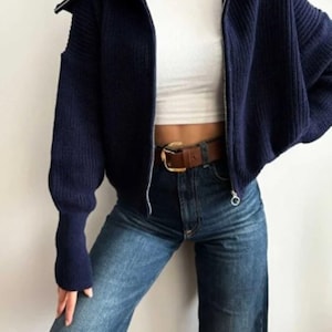 May include: A navy blue zip-up sweater with a wide collar is paired with a white crop top and blue jeans. A brown leather belt with a gold buckle and a gold chain necklace complete the outfit. The sweater has a ribbed texture.