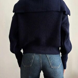 May include: A navy blue sweater with a wide, ribbed collar and long sleeves. The sweater is paired with blue denim jeans. The back view of the outfit is shown against a plain background, highlighting the texture and style of the clothing.