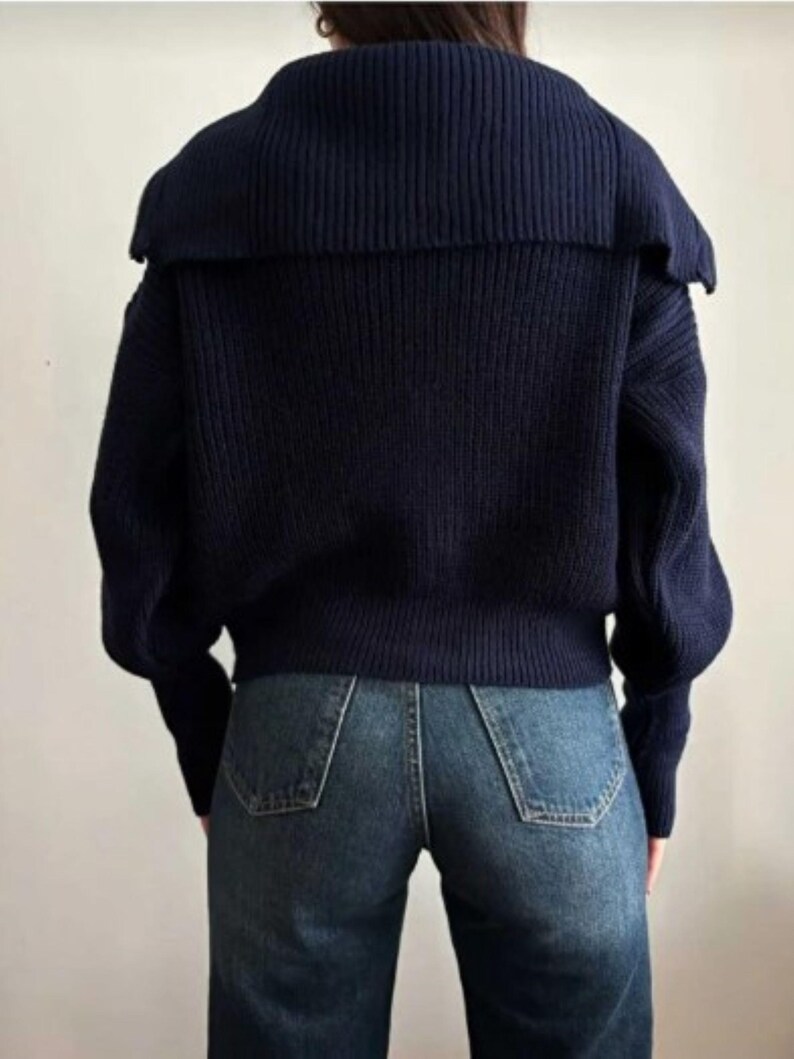 May include: A navy blue sweater with a wide, ribbed collar and long sleeves. The sweater is paired with blue denim jeans. The back view of the outfit is shown against a plain background, highlighting the texture and style of the clothing.