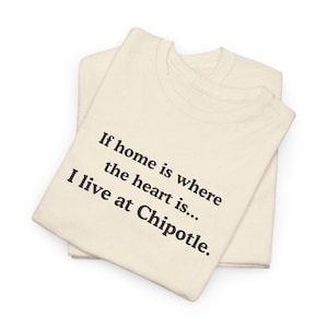 Può includere: Due magliette piegate, color crema. La maglietta superiore ha il testo: "Se la casa è dove è il cuore... Vivo da Chipotle". Il testo è in nero, con un carattere sans-serif. Le magliette sono su sfondo bianco.