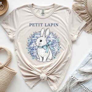 May include: A cream-coloured t-shirt with a white rabbit illustration, blue bow, and floral wreath. The text "PETIT LAPIN" is printed above the rabbit. The shirt has a crew neck and short sleeves.
