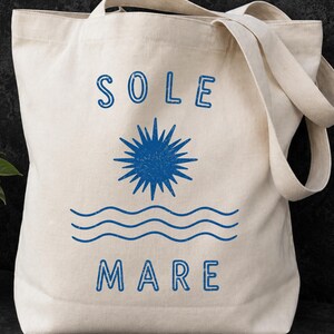May include: A cream-colored canvas tote bag featuring a blue graphic print. The design includes the word SOLE at the top, a stylized sunburst in the center, three wavy lines representing water below the sun, and the word MARE at the bottom.