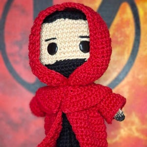 Pode incluir: Boneca de crochê com capa vermelha com capuz, calças e sapatos pretos. A boneca tem um rosto bege com olhos pretos e barba preta. O texto "PDF Pattern" é visível, junto com bandeiras.