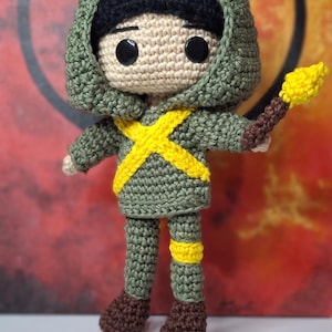 May include: Crocheted doll in a green hooded outfit with a yellow cross design and brown boots. The doll holds a yellow torch. The doll's head is tan with black eyes and a black hood.