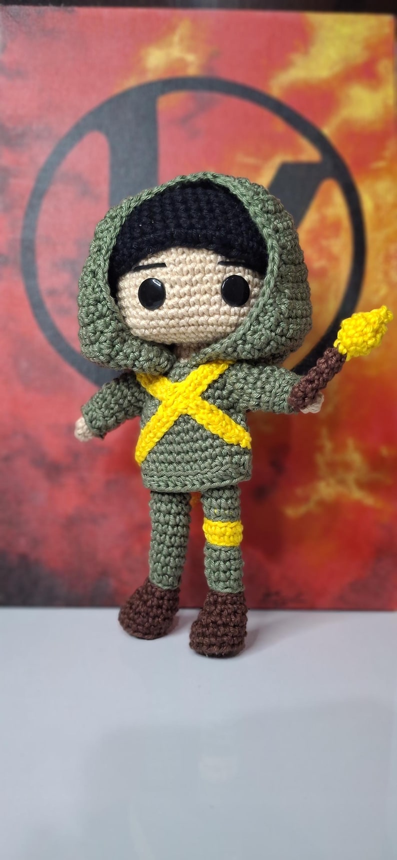 May include: Crocheted doll in a green hooded outfit with a yellow cross design and brown boots. The doll holds a yellow torch. The doll's head is tan with black eyes and a black hood.