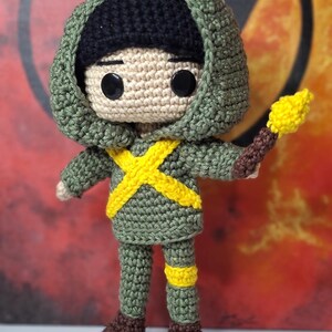 May include: A handmade crochet doll in a green outfit with a black hood and a yellow cross design. The doll holds a brown and yellow torch. The doll's boots are brown. The background is a blurred red and orange.