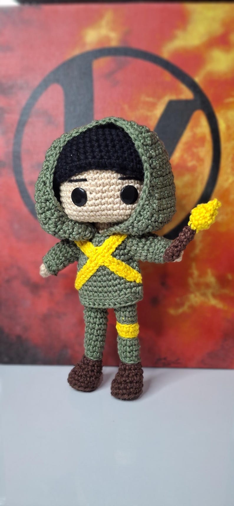 May include: A handmade crochet doll in a green outfit with a black hood and a yellow cross design. The doll holds a brown and yellow torch. The doll's boots are brown. The background is a blurred red and orange.
