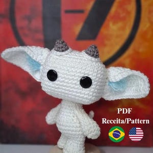 May include: A white crochet creature with large black eyes, gray horns, and large ears with blue accents. The handmade amigurumi stands on a wooden base. Text on the image reads "PDF Receita/Pattern".
