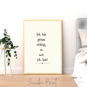 May include: A framed print with the text "Ich bin genau richtig, so wie ich bin!" in black font. The frame is a light wood color, and the background is off-white. A small black heart is below the text. The print is displayed in a bedroom setting.