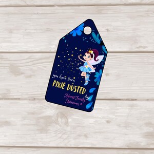 May include: A navy blue gift tag with a cartoon fairy illustration and the text "you have been PIXIE DUSTED". The fairy has a blue dress, wings, and a crown. Decorative blue floral elements surround the edges of the tag.