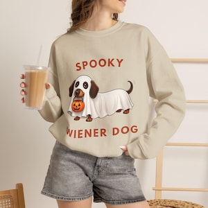Puede incluir: Una sudadera beige de manga larga con el gráfico de un perro salchicha vestido con un disfraz de fantasma blanco y sosteniendo un pequeño cubo de calabaza naranja. El texto SPOOKY aparece arriba y WIENER DOG abajo en color rojo.
