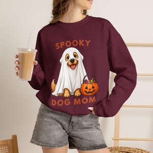 Puede incluir: Una sudadera de cuello redondo y manga larga color granate con un gráfico de un golden retriever vestido con un disfraz de fantasma blanco. El perro sostiene una cesta de dulces de calabaza. El texto dice SPOOKY DOG MOM en letras naranjas.