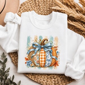 Könnte beinhalten: Ein weißes Langarm-Sweatshirt mit einer zentralen Grafik eines Kürbisses, dessen Segmente verschiedene Muster wie Leopardenmuster, orangefarbenes Vichy-Karo und blaue Blumen aufweisen, verziert mit einer großen blauen Schleife und Herbstblättern.