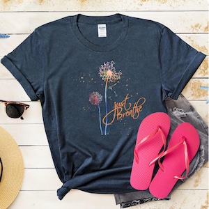 Pode incluir: Camiseta azul marinho com um desenho de dente-de-leão e a frase "just Breathe" em laranja. A imagem também mostra chinelos rosa, óculos de sol e um chapéu de palha. É uma peça de roupa casual.