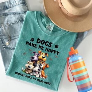 May include: A teal t-shirt with the text "DOGS MAKE ME HAPPY" and a graphic of six cartoon dogs. A beige hat, blue jeans, a water bottle with orange and blue stripes, and a necklace are also in the image.