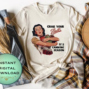 May include: A cream-coloured t-shirt with a vintage illustration of a woman holding jars of preserves. The text "Grab Your Balls, It's Canning Season" is printed in red and black. The shirt is part of a digital download.