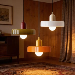 Può includere: Tre moderne lampade a sospensione rosse, verde oliva e arancioni. Ogni lampada ha un paralume circolare e una lampadina sferica. Le lampade pendono da cavi neri contro una parete a pannelli di legno e un tappeto fantasia.
