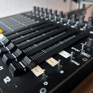 Puede incluir: Primer plano de una consola de mezcla de audio negra con múltiples deslizadores, perillas y botones. La consola tiene etiquetas como "NOTES", "FALSE", "ARM" y "REC". Los deslizadores están dispuestos en filas paralelas y las perillas están en la parte superior.