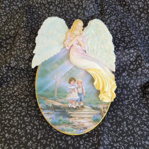 May include: An oval wall plaque featuring a three-dimensional angel with large cream-colored wings and a flowing gown. The center depicts a painted scene of two children standing on a wooden bridge over a stream, surrounded by flowers and trees.