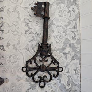 May include: A decorative wall-mounted key featuring a dark bronze finish with copper-toned highlights. The key has a long cylindrical shaft and a large, ornate bow with intricate scrollwork and a central circular medallion detail.