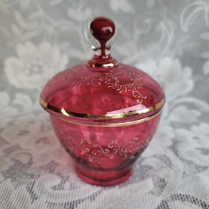 May include: A deep cranberry red glass lidded jar featuring a gold rim and delicate gold filigree scrollwork patterns on both the bowl and the lid. The lid is topped with a clear glass finial containing a small red sphere at its center.