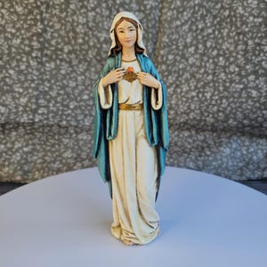 May include: A standing figurine of the Virgin Mary wearing a cream-colored robe and a teal blue mantle. The figure has a veil covering the head and hands crossed over the chest, where a small, detailed golden heart emblem is visible.