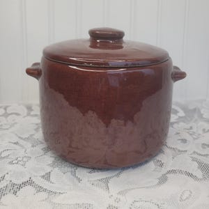 May include: A cylindrical brown ceramic pot with a matching lid and two small side handles. The exterior features a smooth, glossy finish with a deep, solid brown color throughout the body and the lid, which has a small knob on top.