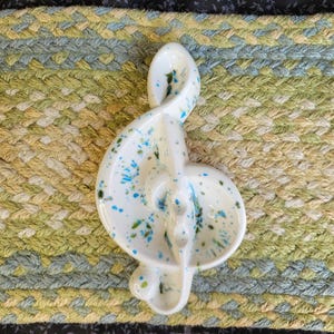 May include: A white ceramic treble clef shaped dish featuring a glossy finish with a speckled pattern of bright blue and green paint splatters across the surface. The musical symbol design creates a curved, concave shape suitable for holding small items.