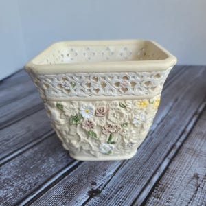 May include: Cream-colored square ceramic planter with a heart-shaped lattice design around the rim. The planter features embossed floral and leaf details in white, yellow, and pink. The planter is designed for indoor or outdoor use.