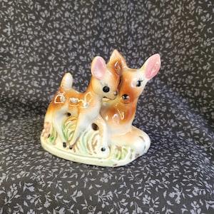 May include: A ceramic figurine featuring two deer standing together on a base painted with green grass. The deer are colored in shades of tan, brown, and white with black eyes and pink-tinted ears. The piece has a glossy finish throughout.